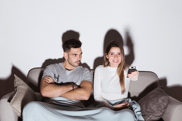 Woman changing TV channels while man looks annoyed on couch