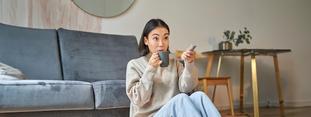 Young Korean woman watching television with remote control looking amazed